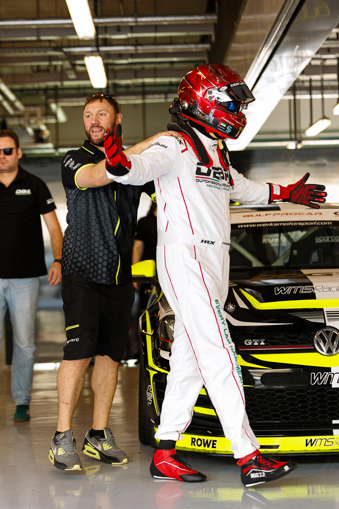 A Dutch Brothers Racing driver in a white race suit and red helmet having his gloves checked by a crew member in the garage.