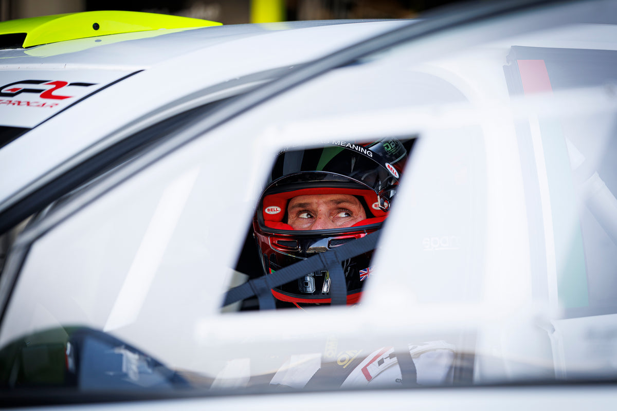 The driver belted into the cockpit in his red helmet, seen through the side window and roll cage.