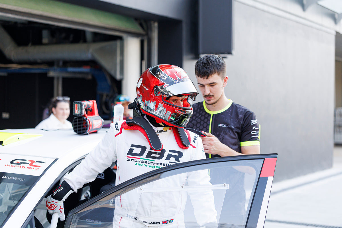 A Dutch Brothers Racing driver in his red helmet climbing into the car, a crew member holding the door open.