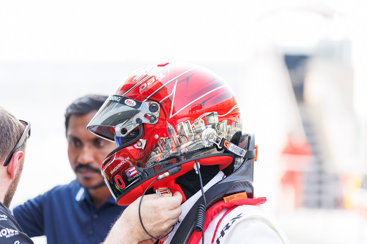 A crew member fastening a driver's red helmet in the pit lane.