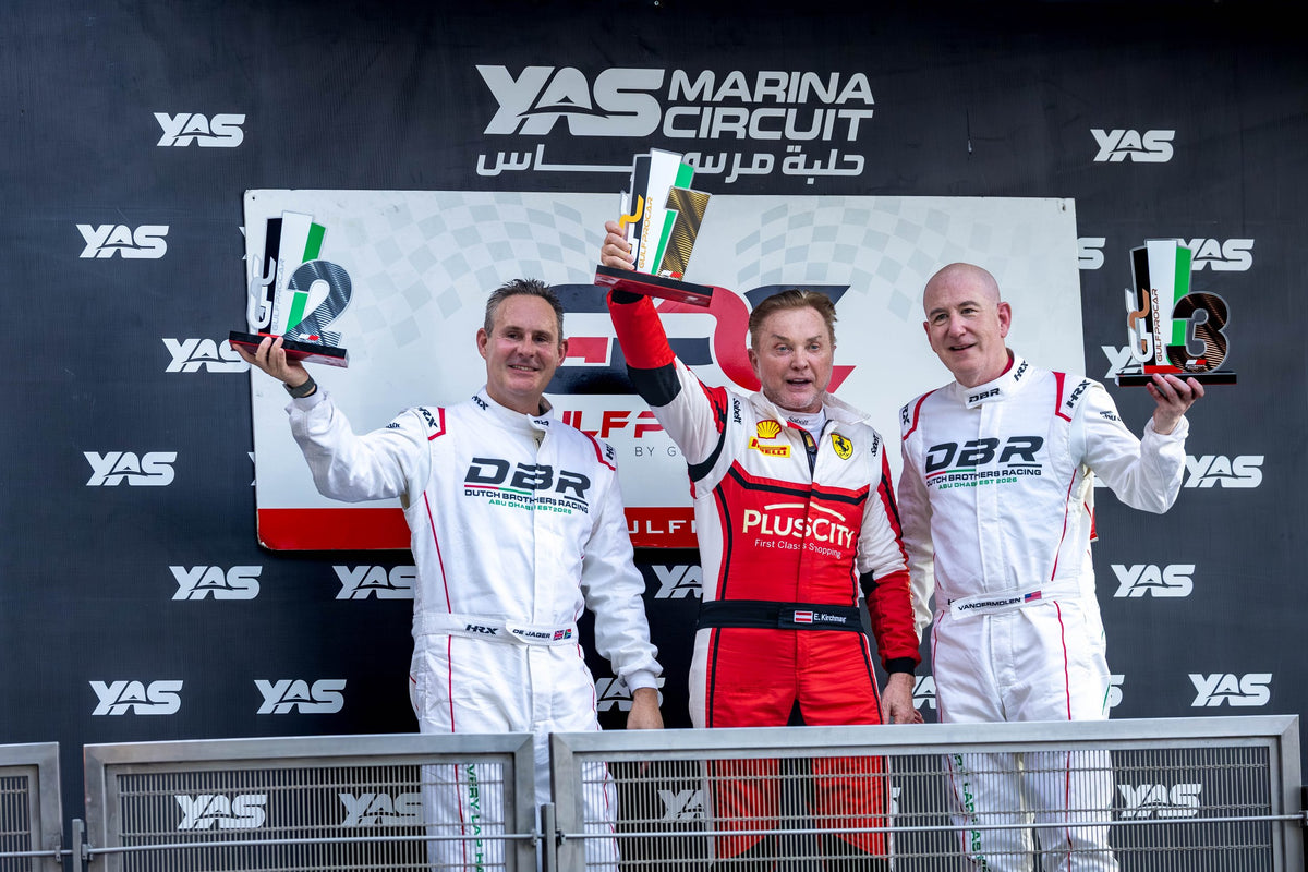 Jon de Jager and Alan VanderMolen on the second and third steps of the podium at Yas Marina Circuit in their white Dutch Brothers Racing suits, trophies raised, the race winner between them in red.